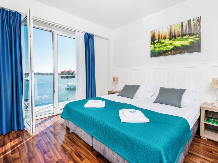 A hotel room with a large bed, a view of the sea through a sliding door, and a painting of a forest on the wall.