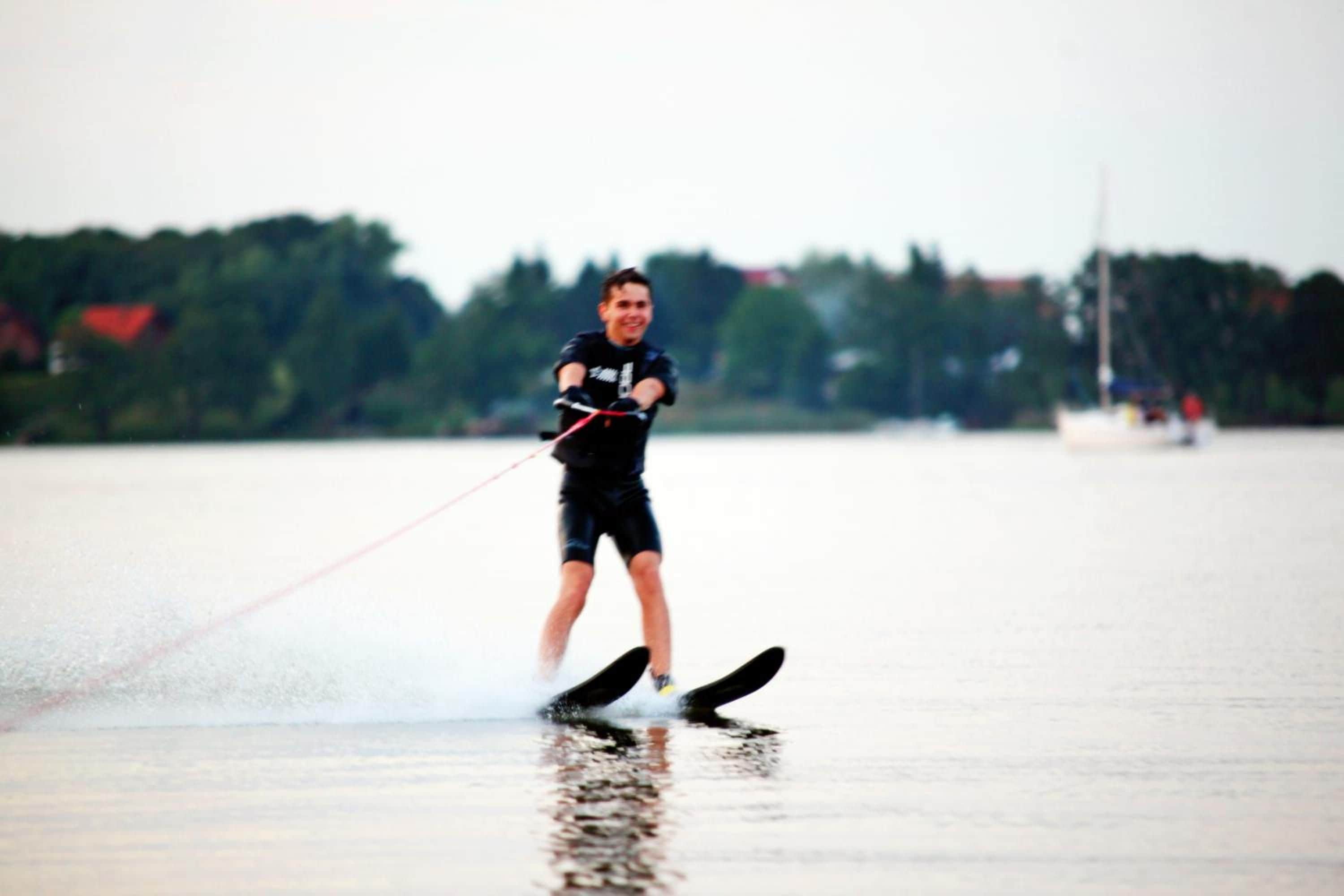 Una persona practicando wakeboard sobre aguas tranquilas, con un fondo borroso de árboles y edificios.