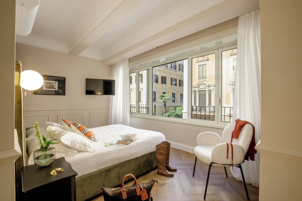 A hotel room with a bed, a chair, a desk with a lamp, a window with a view of buildings, and a vase with flowers on the nightstand.
