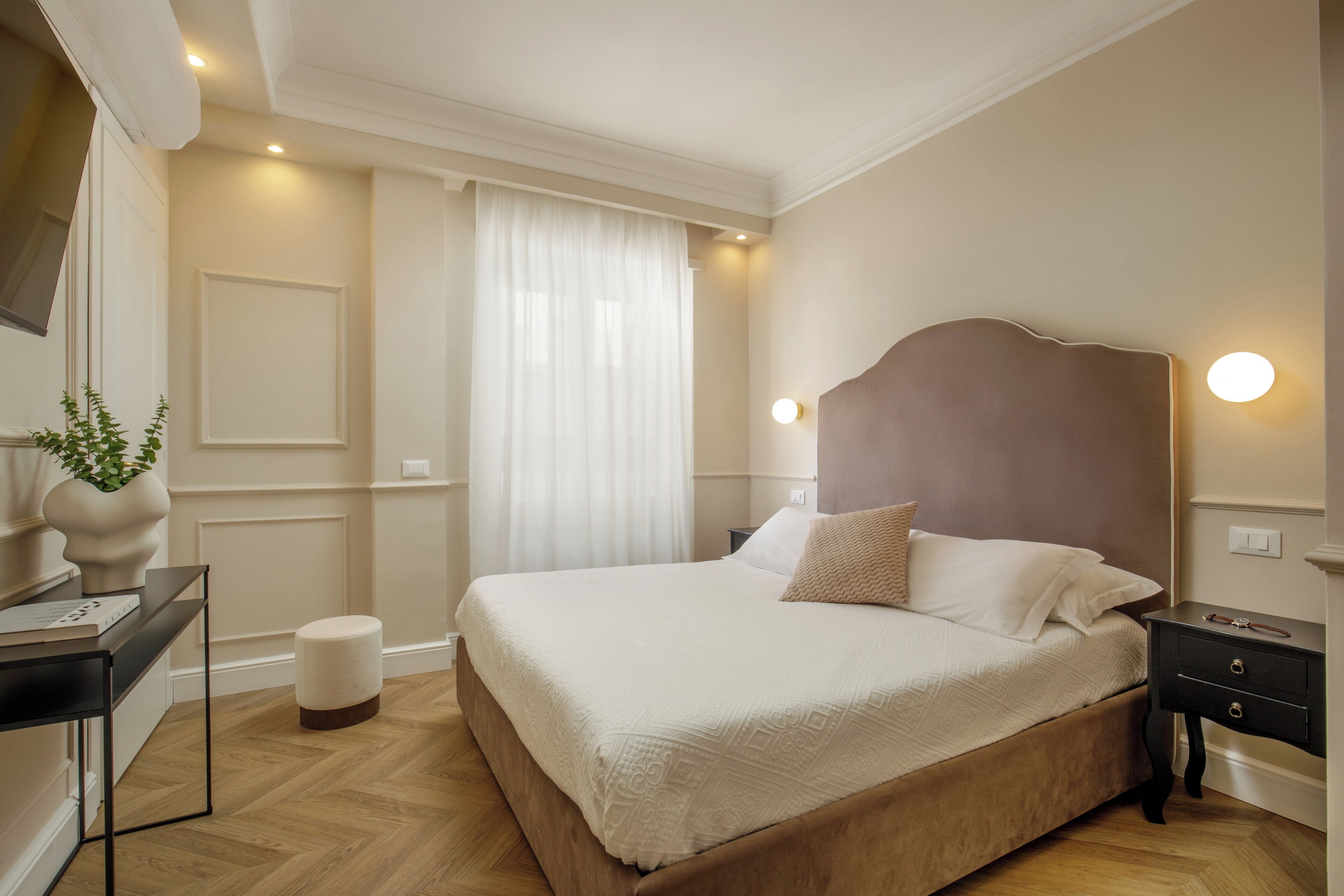 A neatly arranged bedroom with a large bed, bedside tables, a small stool, and a potted plant.