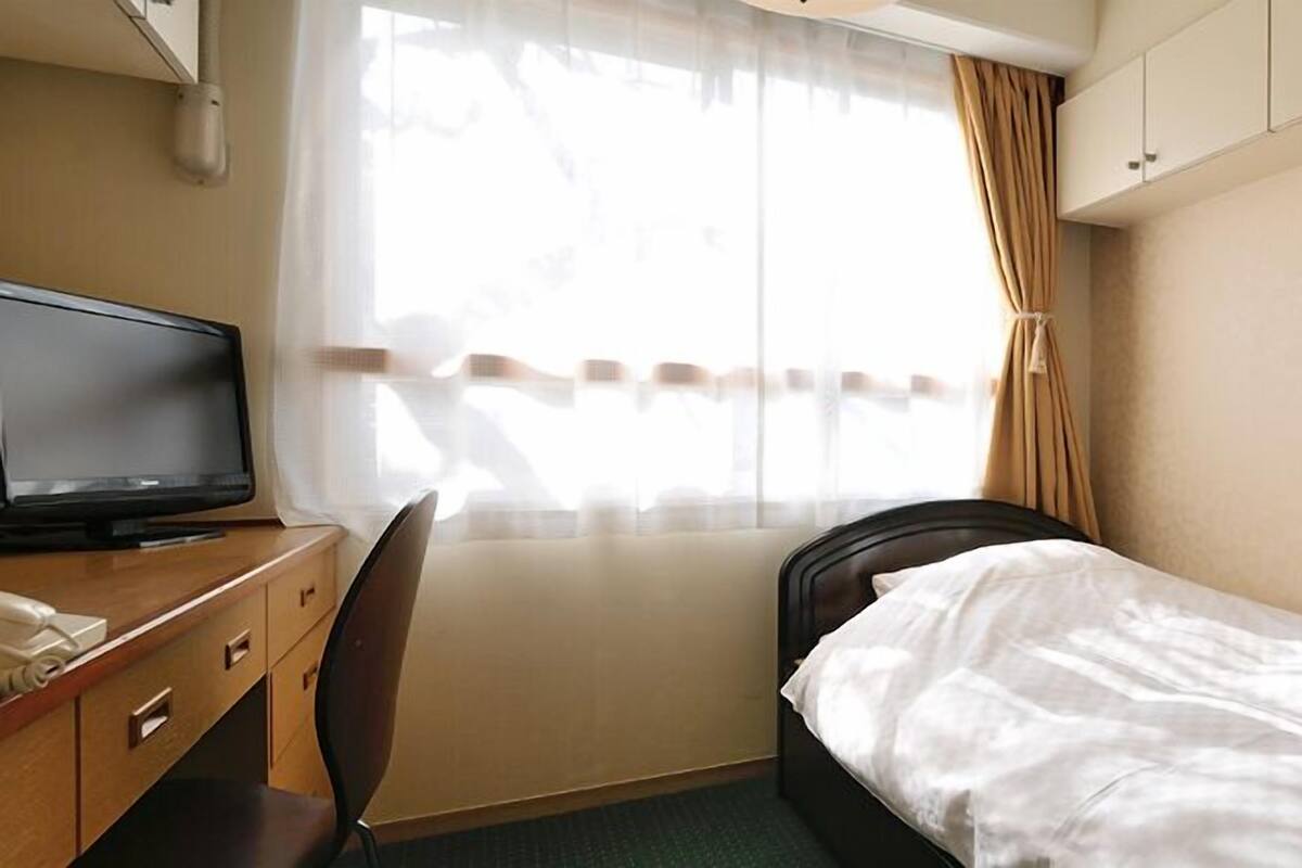 Habitación de hotel con cama, escritorio con televisor, silla y ventana con cortinas.