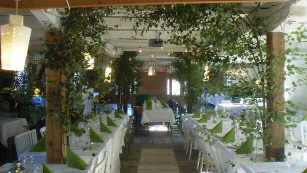 A long dining table with white tablecloths and green napkins, surrounded by white chairs, under a wooden pergola with hanging lights. - Brunnsta Gård (Balsta)