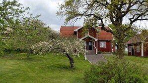 A red house with a red roof, surrounded by green grass and trees with pink blossoms. - Brunnsta Gård (Balsta)
