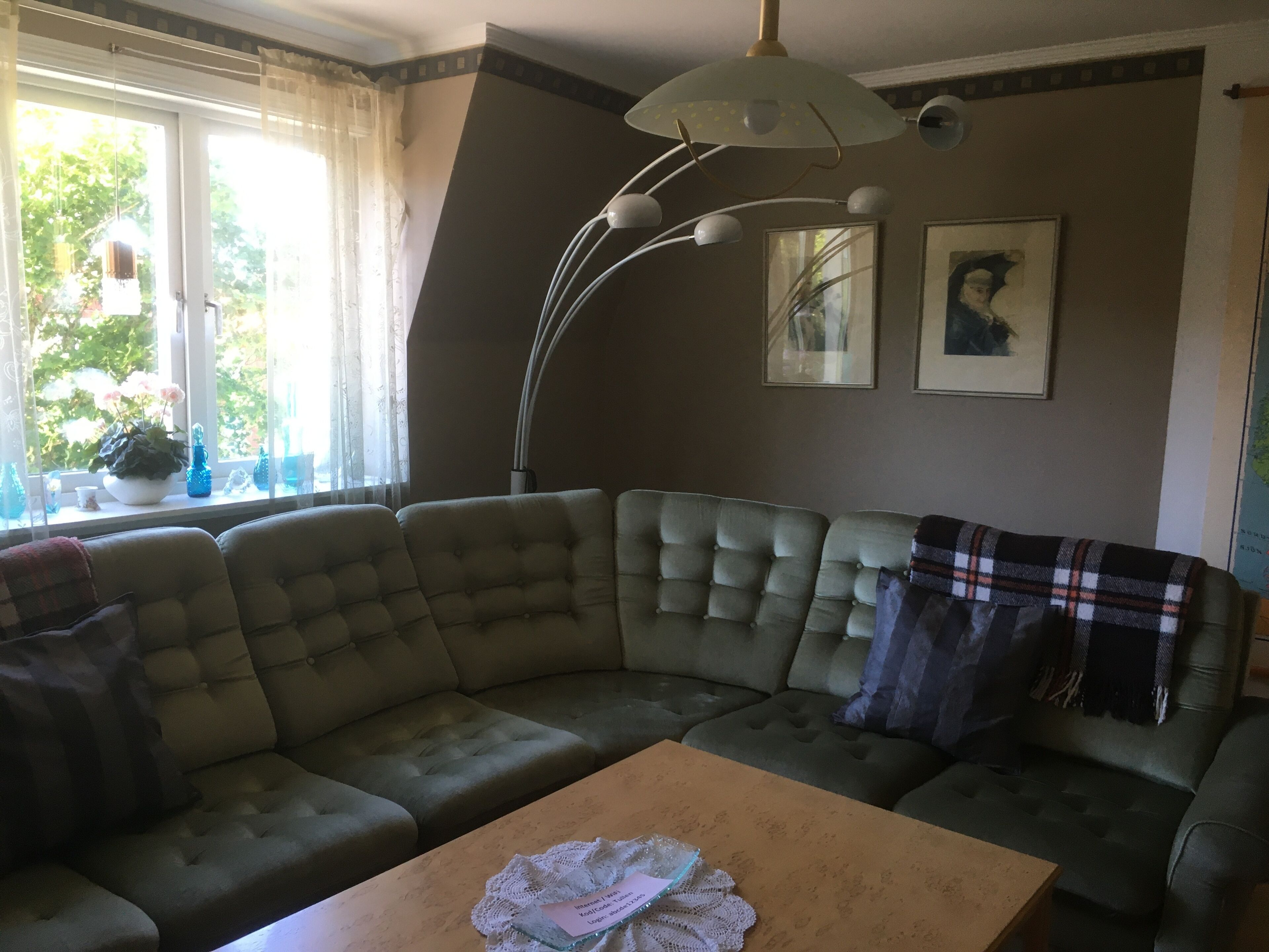 A living room with a green sofa, a wooden coffee table, and a floor lamp. There are two framed pictures on the wall and a window with curtains.