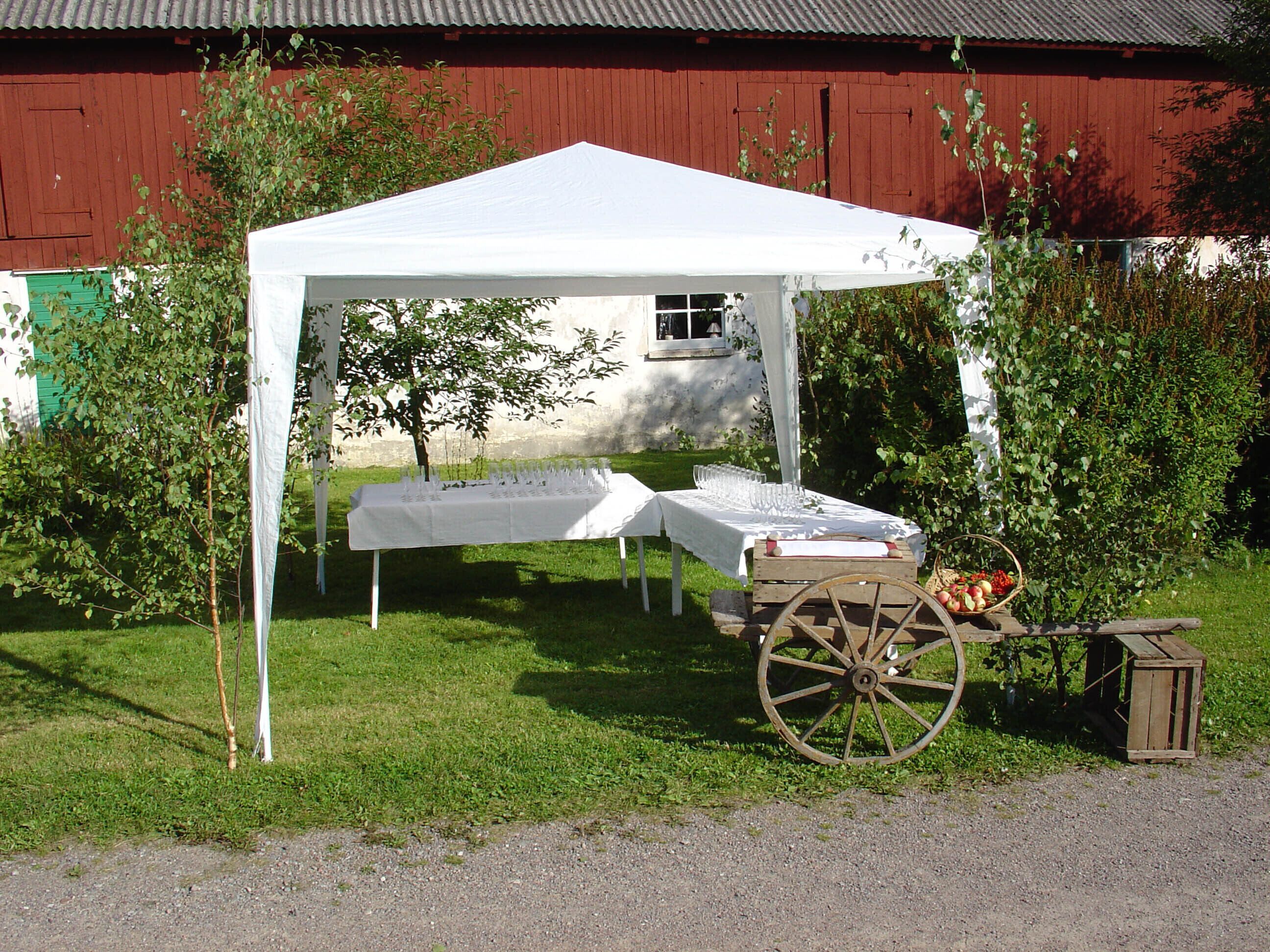 A white tent set up outdoors with a table covered in a white tablecloth, a wooden cart with a basket of fruit, and a red barn in the background.