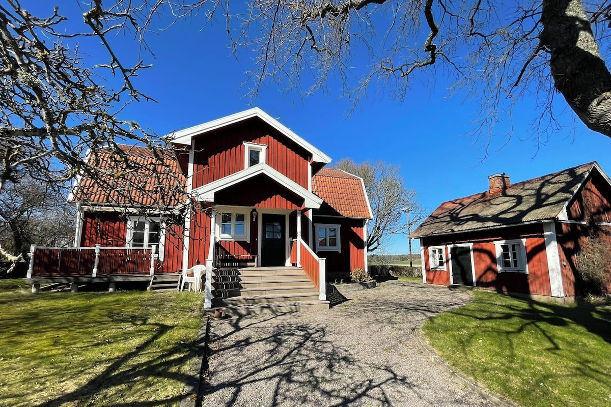 A red wooden house with a porch, a small white chair, and a gravel path leading to the entrance.