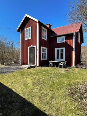 A two-story red wooden house with a red roof, a front door, and a small patio with two chairs. - Brunnsta Gård (Balsta)