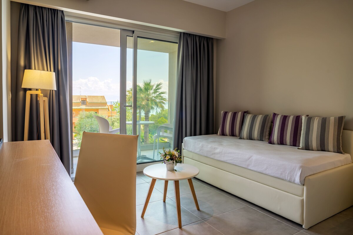 A modern hotel room with a sofa, a small round table with a lamp, and a view of palm trees through the glass door.