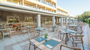 An outdoor cafe area with wicker tables and chairs, a large building with multiple balconies, and a clear sky. - CNic Gemini Hotel (Corfu)