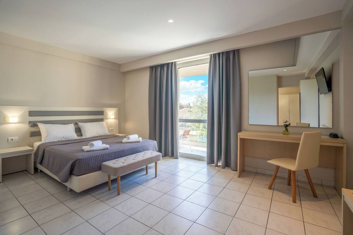 A modern hotel room with a bed, bedside tables, a desk, a chair, and a balcony with curtains.