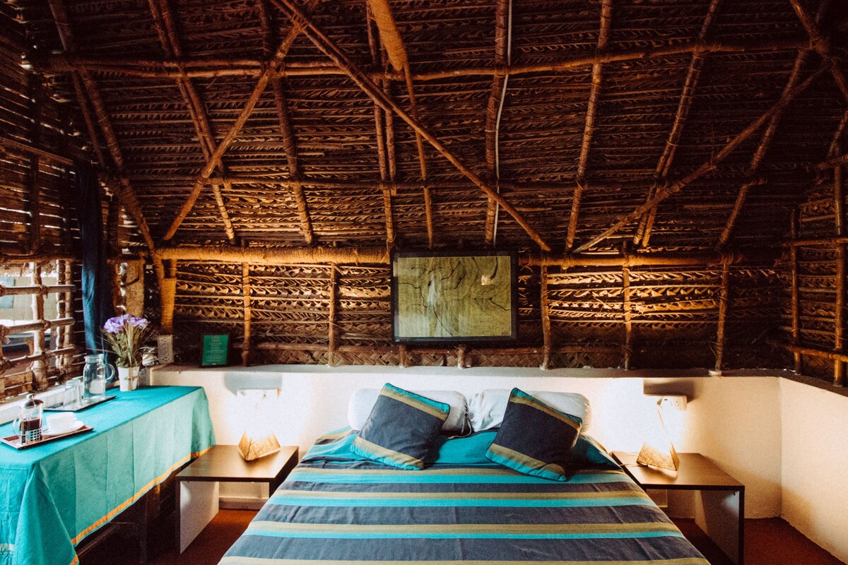 Um quarto com telhado de palha, uma cama com roupa de cama listrada, uma pequena mesa com um vaso de flores e um quadro na parede.