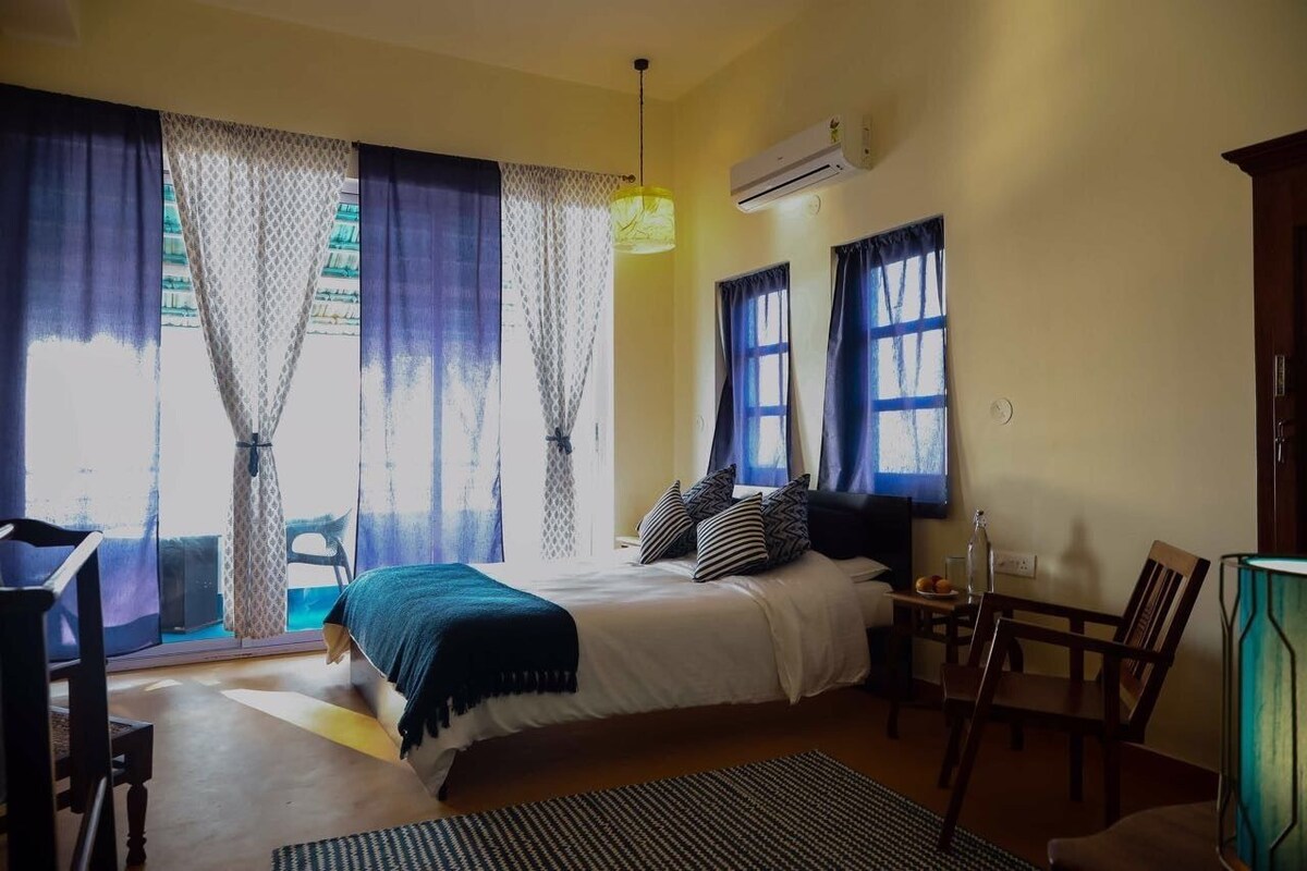 Quarto com cama, escrivaninha e cadeira. Há uma janela com cortinas e um ar condicionado na parede.