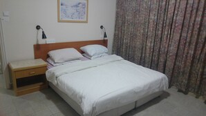 A neatly made bed with white bedding, a wooden nightstand with a drawer, two wall-mounted lamps, and a painting above the bed. - Marom Ziv Center - Newe Shaanan (Haifa)