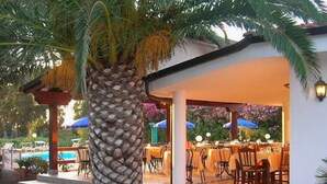 An outdoor dining area with tables and chairs, a palm tree, and a pool in the background. - C'era Una Volta (Ricadi)