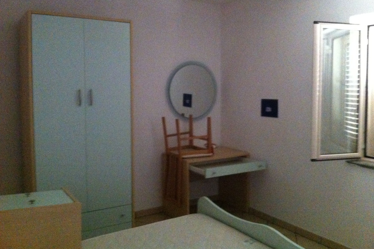 A bedroom with a bed, a wardrobe, a small table with a mirror, and a window with blinds.
