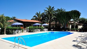 A swimming pool with lounge chairs and umbrellas, surrounded by palm trees and a building with a red roof. - C'era Una Volta (Ricadi)