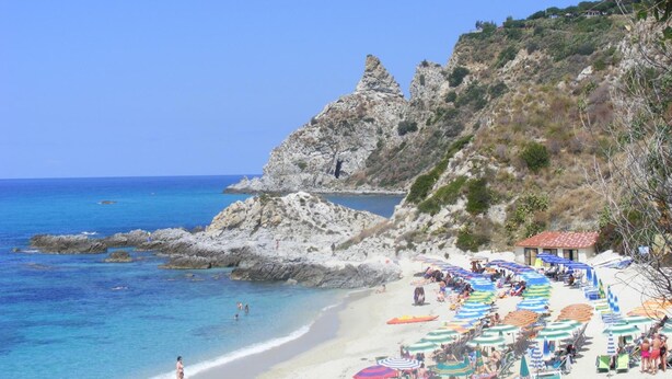 A beach with rocky cliffs, clear blue water, and numerous colorful umbrellas and sun loungers. - C'era Una Volta (Ricadi)