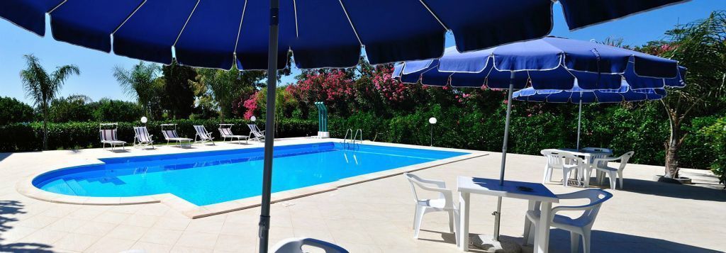 Une piscine entourée de verdure, avec des chaises longues et des parasols, sous un ciel dégagé.