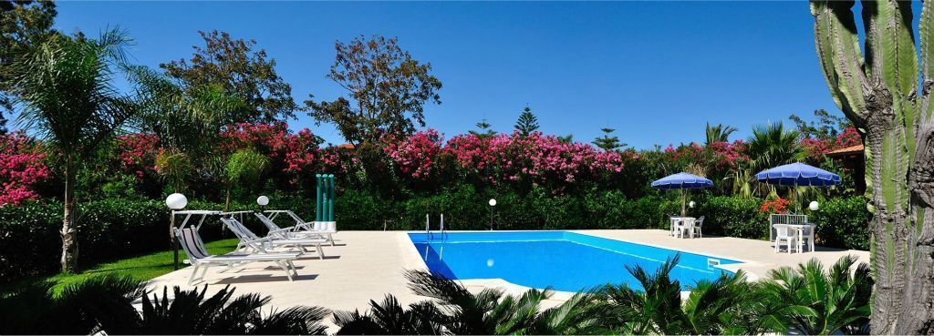 Una piscina con lettini e ombrelloni, immersa nel verde e circondata da piante fiorite.