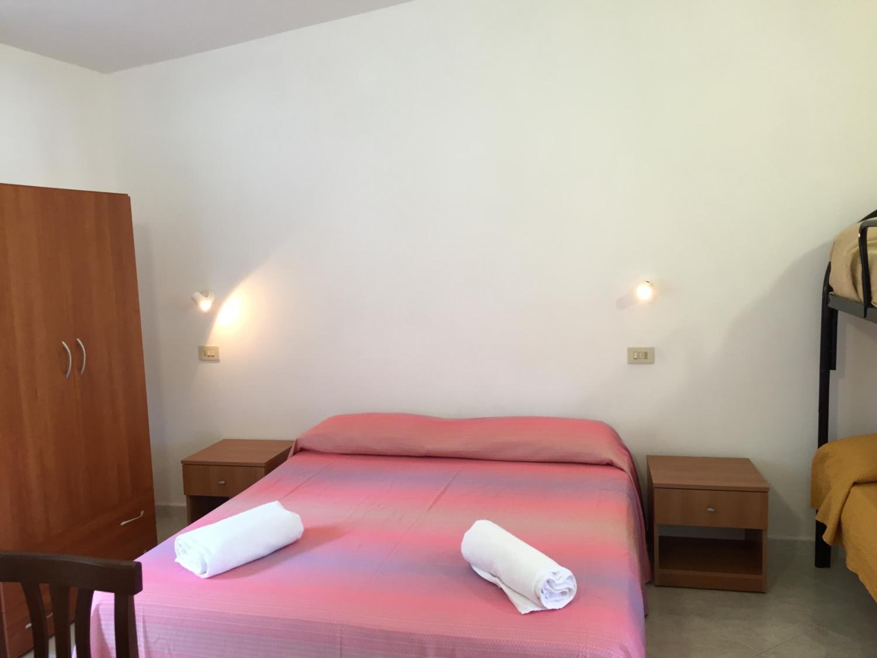 A single bed with a pink blanket, two bedside tables, a wooden wardrobe, and two wall-mounted lights.