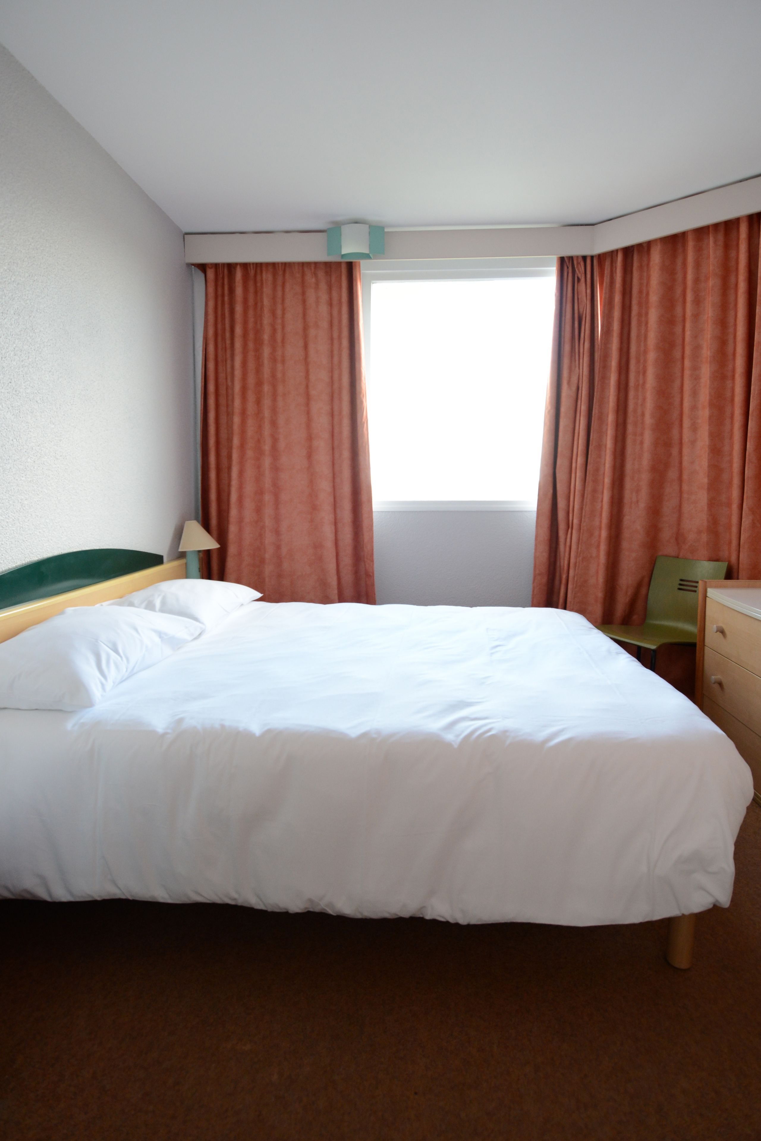 A hotel room with a bed, a chair, a dresser, and a window with curtains.