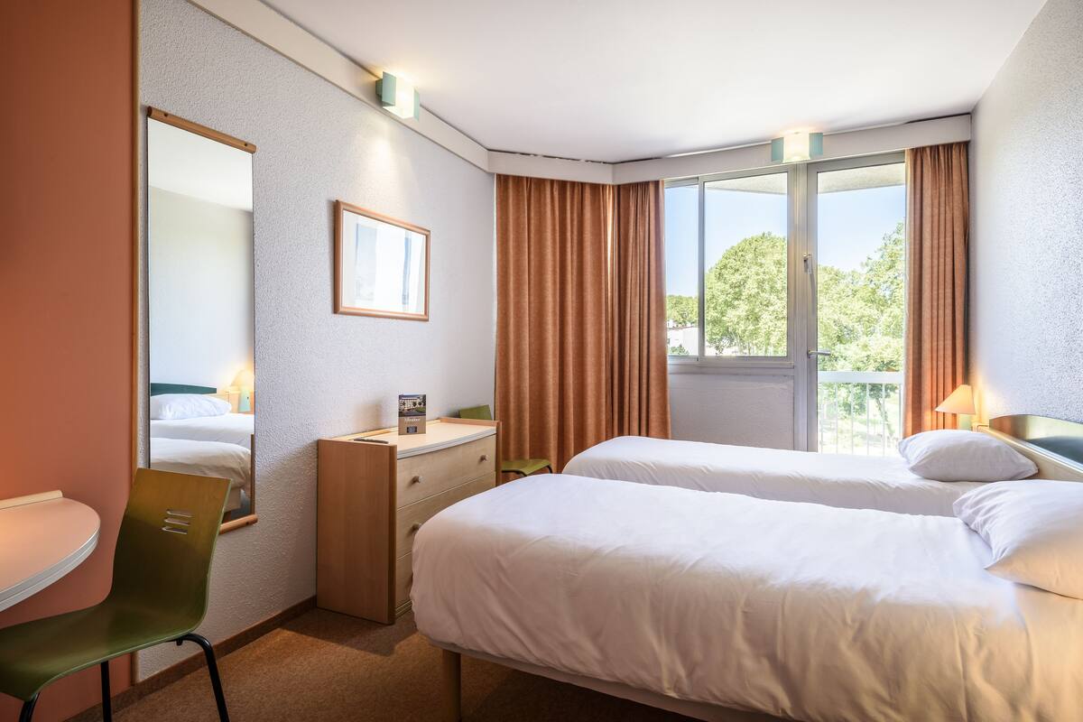 Ein Hotelzimmer mit zwei Betten, einem Schreibtisch, einem Sessel, einer Kommode und einem großen Fenster mit Vorhängen.