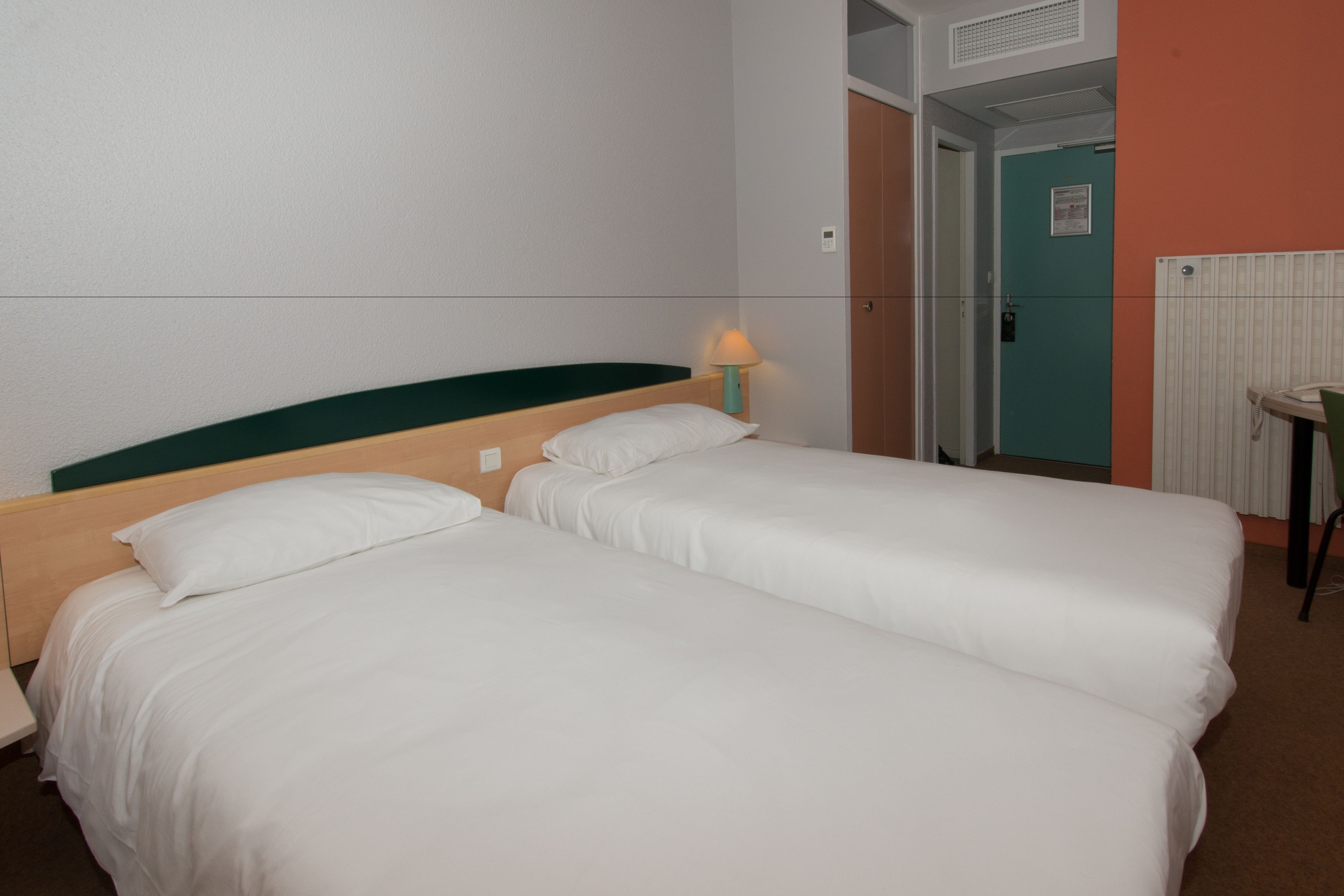 Habitación de hotel con dos camas individuales, una mesita de noche, una lámpara y una puerta que da a un baño.