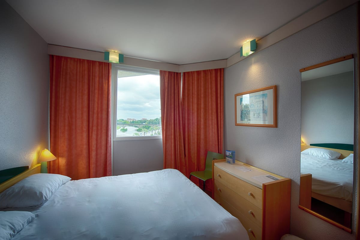 Ein Hotelzimmer mit einem Bett, einem Schreibtisch, einem Stuhl, einer Kommode, einem Fenster mit Vorhängen, einem Bild und einem Spiegel.