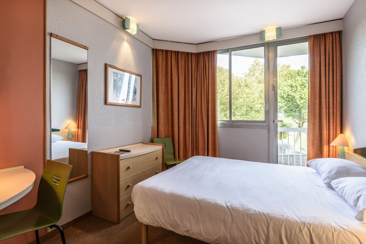 Ein Hotelzimmer mit Bett, Schreibtisch, Stuhl, Kommode, Spiegel und einem Fenster mit Vorhängen.