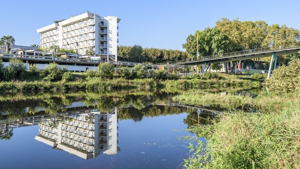 A modern hotel with a reflection in a calm water body, surrounded by greenery and a pedestrian bridge. - Hôtel Miradour (Dax)