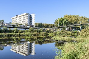Un hotel moderno con su reflejo en un cuerpo de agua tranquilo, rodeado de follaje y un puente peatonal.