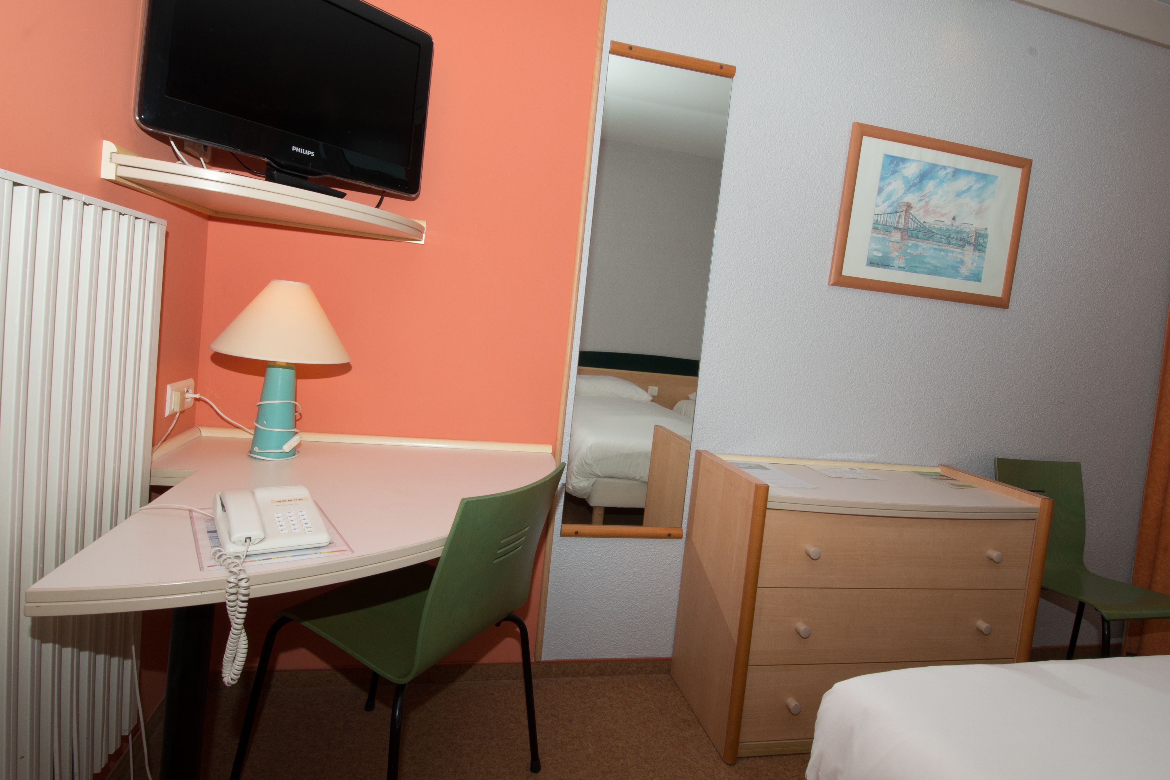 Habitació d'hotel amb un llit, una taula de treball amb una làmpada, una cadira, un calaixer, un televisor i un quadre a la paret.