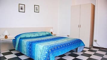 Ein Schlafzimmer mit Schachbrettfußboden, einem Bett mit blaugrün gemustertem Bettbezug, zwei gerahmten Bildern an der Wand und einem weißen Kleiderschrank.