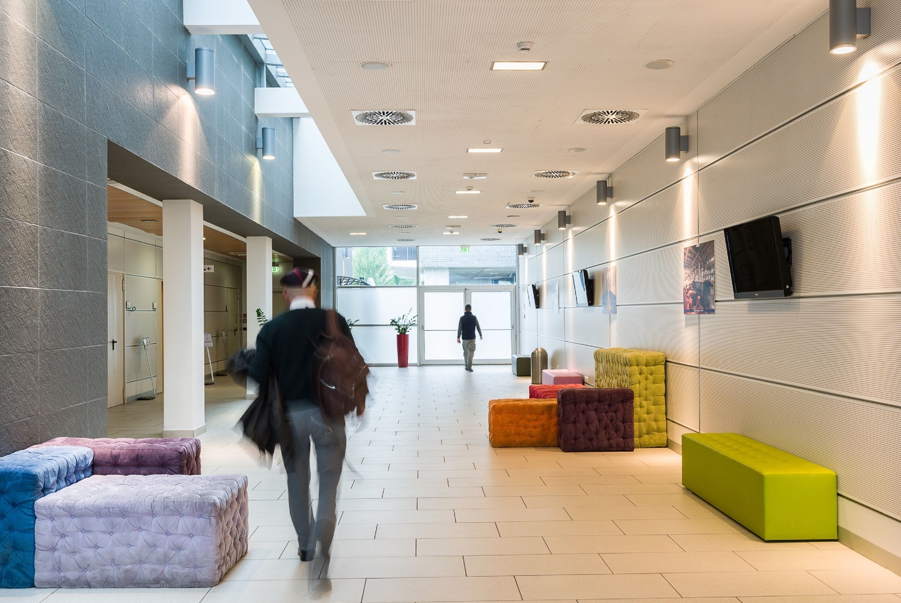 Een modern interieur met een centrale gang, zithoeken en een persoon die naar de uitgang loopt.