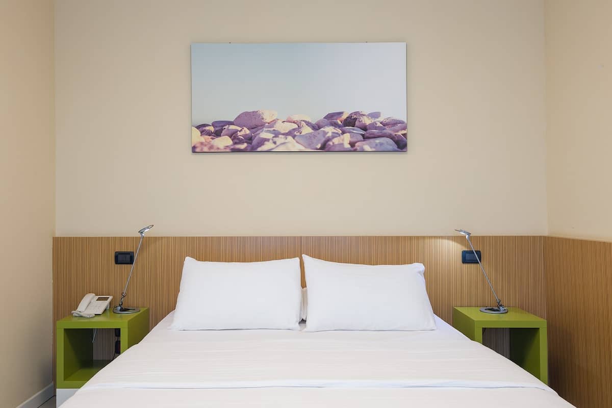 Un lit bien fait, avec du linge de lit blanc, deux tables de chevet avec des lampes et un tableau représentant des rochers au mur.