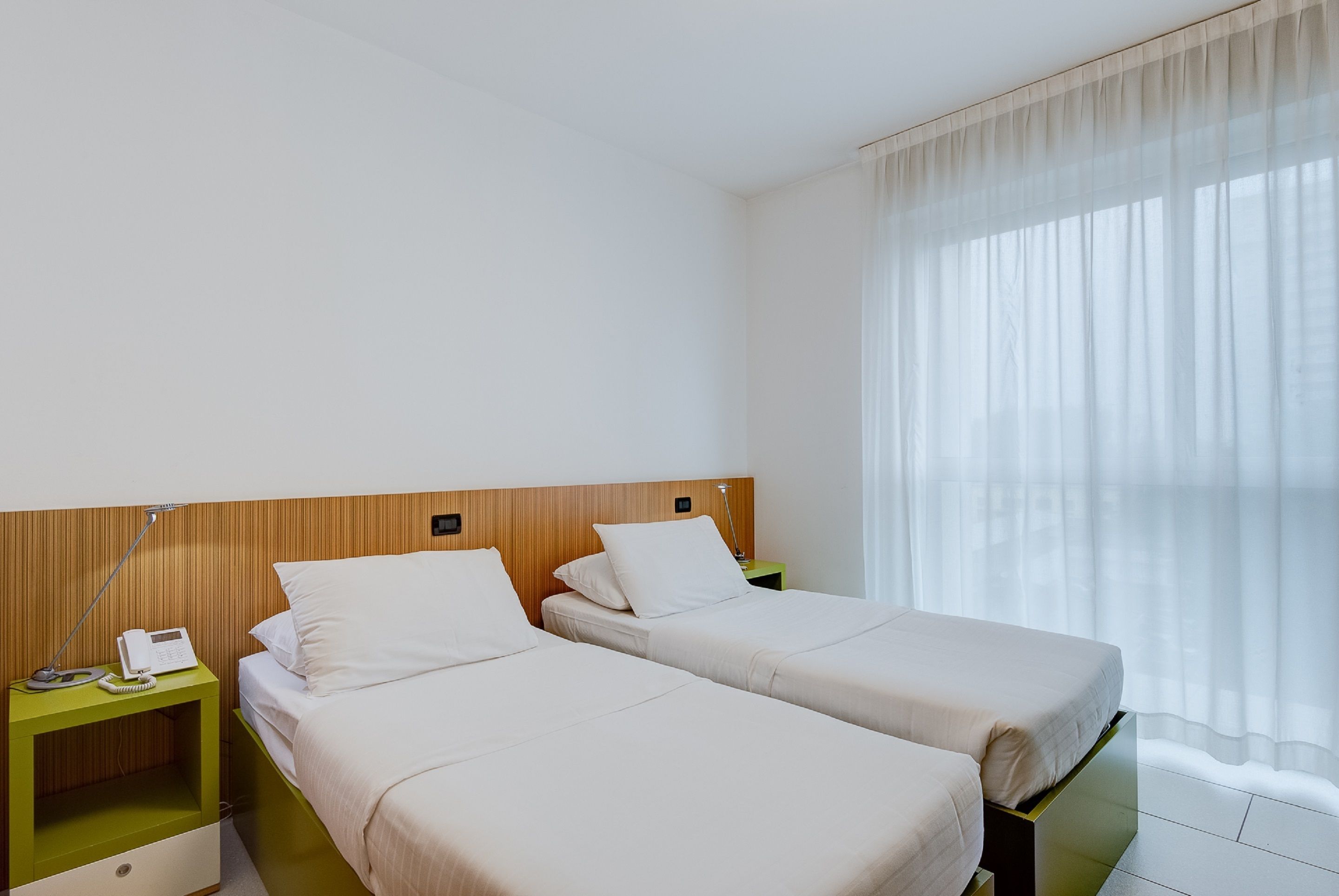 Habitación de hotel con dos camas individuales, cabecero de madera, mesita de noche con lámpara y teléfono.