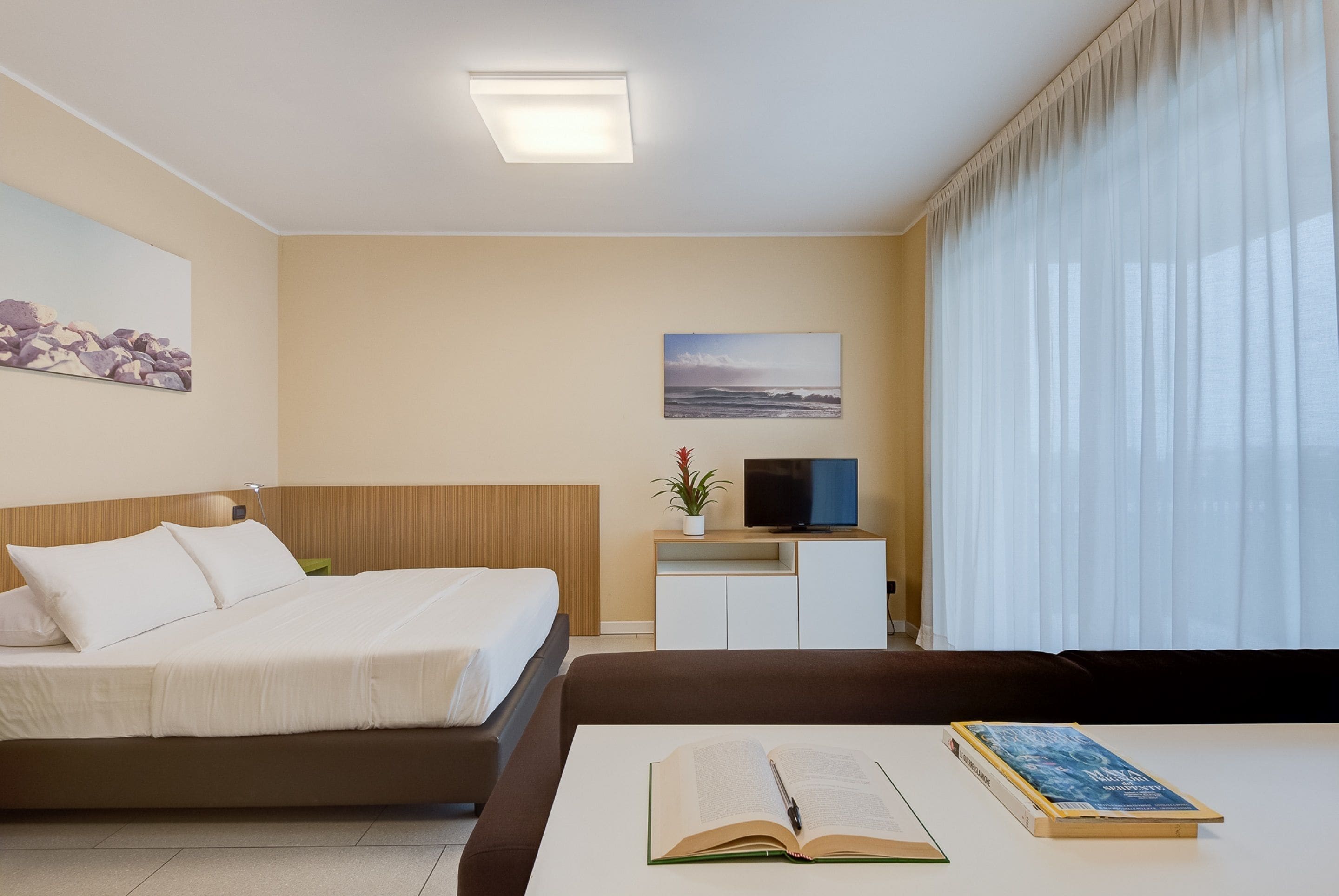 Een hotelkamer met een bed, een televisie op een standaard, een bureau met een boek en een raam met gordijnen.