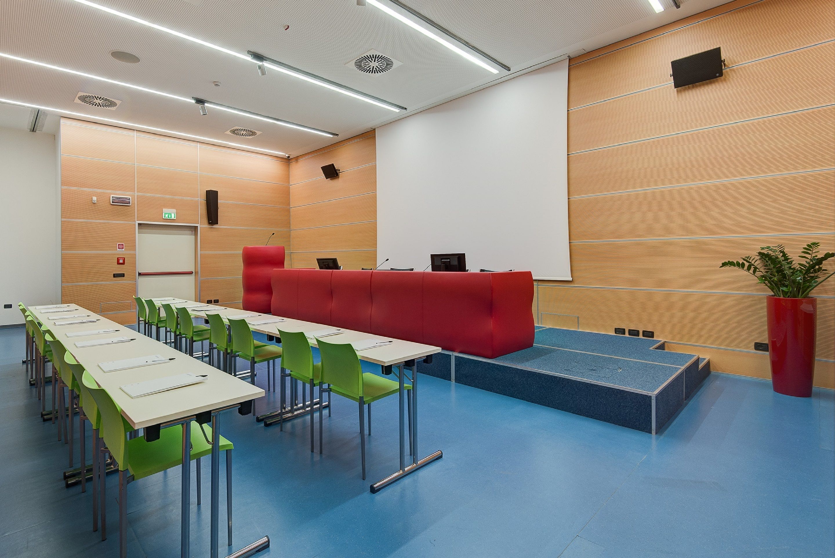 Une salle de conférence avec une longue table, des chaises vertes, un canapé rouge et un écran de projection.