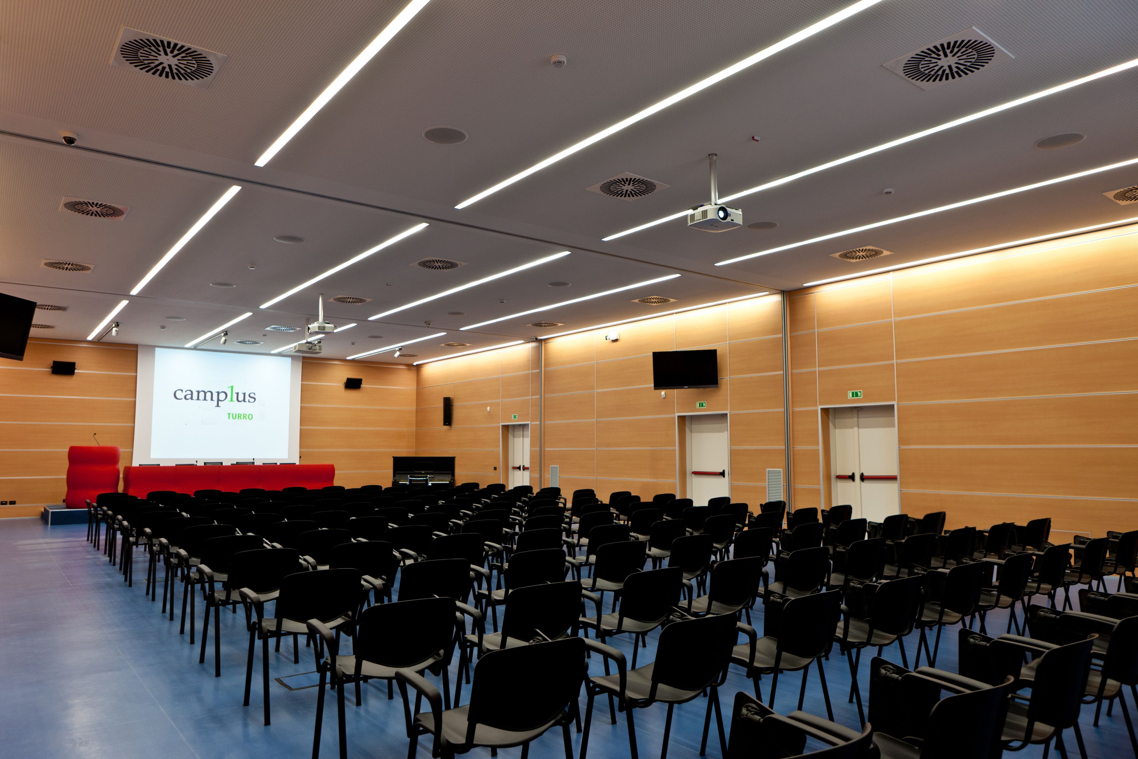 A conference room with rows of chairs, a projector screen, and a red stage.