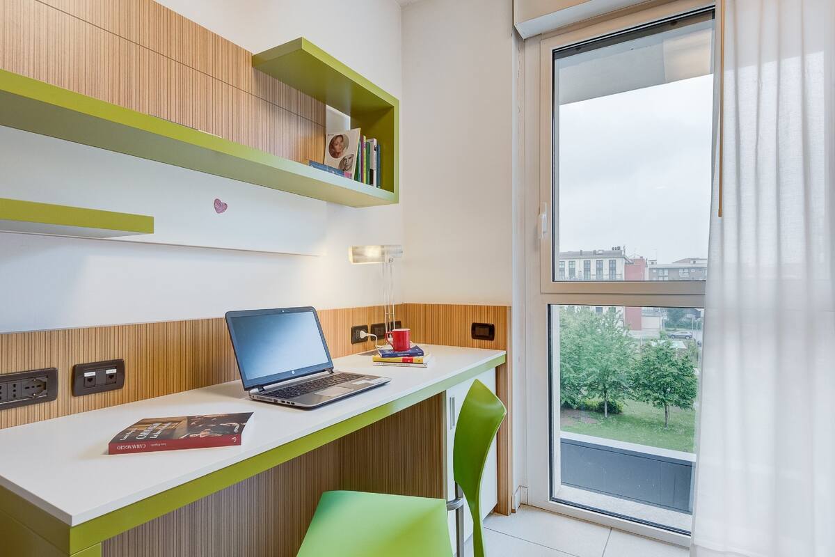Une pièce moderne avec un bureau blanc, un ordinateur portable, une tasse rouge et une chaise verte. Les murs sont recouverts de panneaux en bois et une fen être offre une vue sur des immeubles et des arbres.