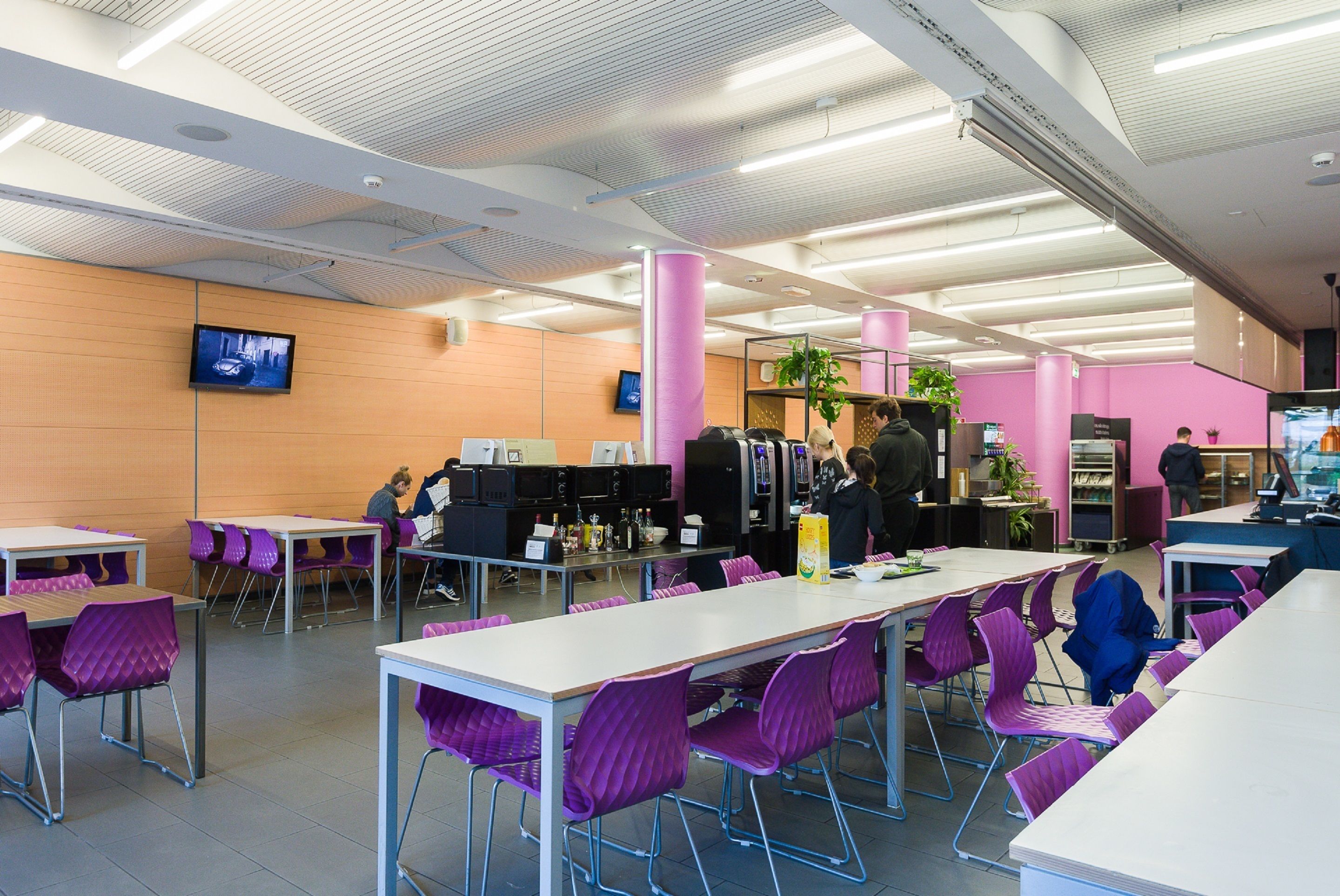 A cafeteria with purple chairs, white tables, and a television on the wall.