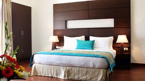 A hotel room with a large bed, two bedside lamps, a wooden headboard, and a floral arrangement on the floor. - Stately Suites MG Road (Gurugram)