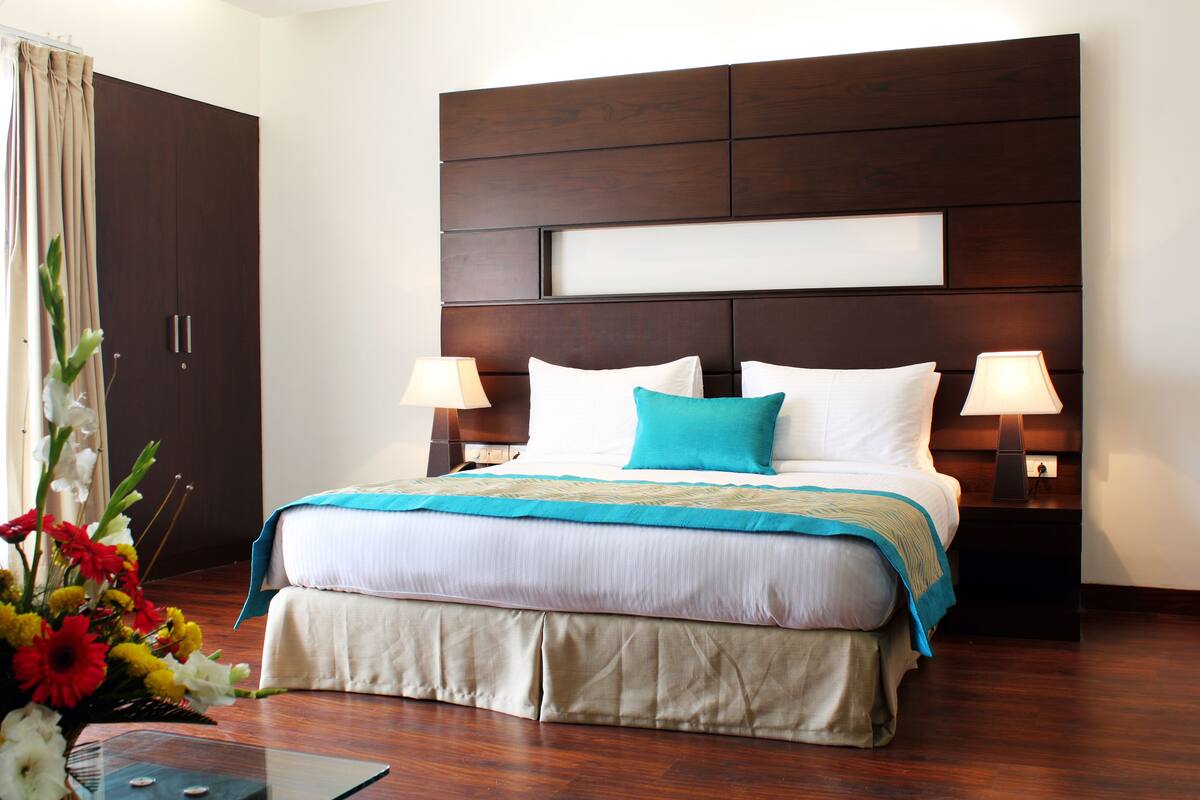 Habitación de hotel con una cama grande, dos lámparas de noche, un cabecero de madera y un arreglo floral en el suelo.