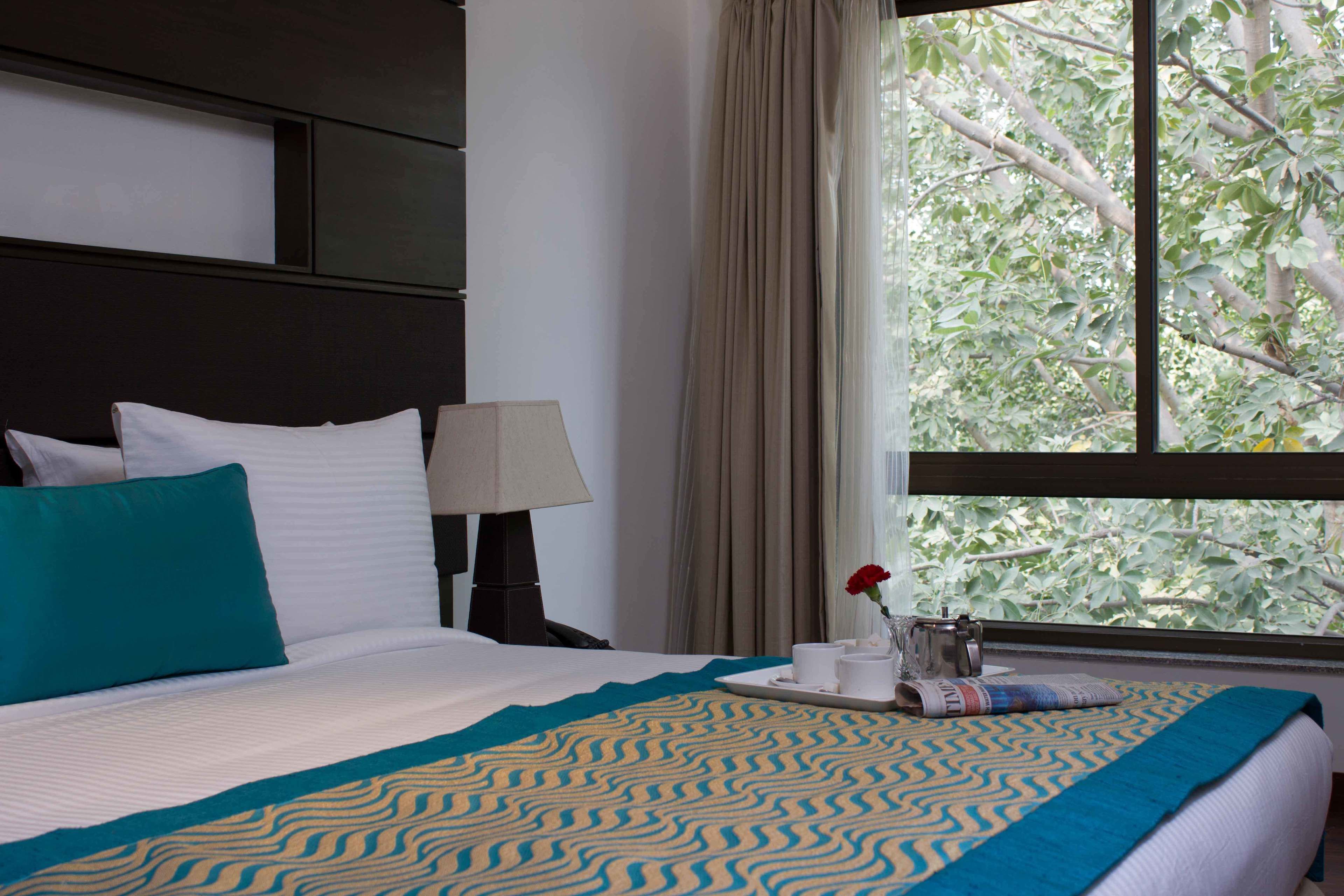 A hotel room with a bed, a nightstand, a lamp, a window with curtains, and a view of trees.