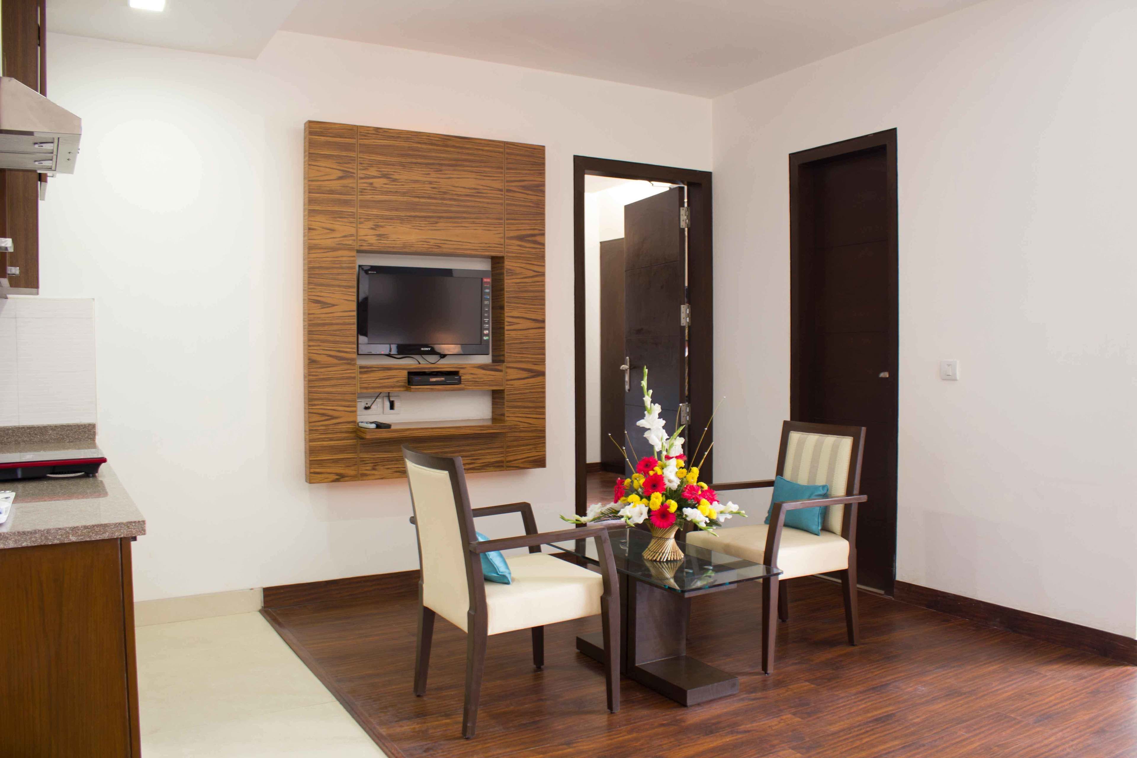 A modern living room with a flat-screen TV mounted on a wooden panel wall, two chairs with cushions, a glass coffee table, and a door with a dark frame.