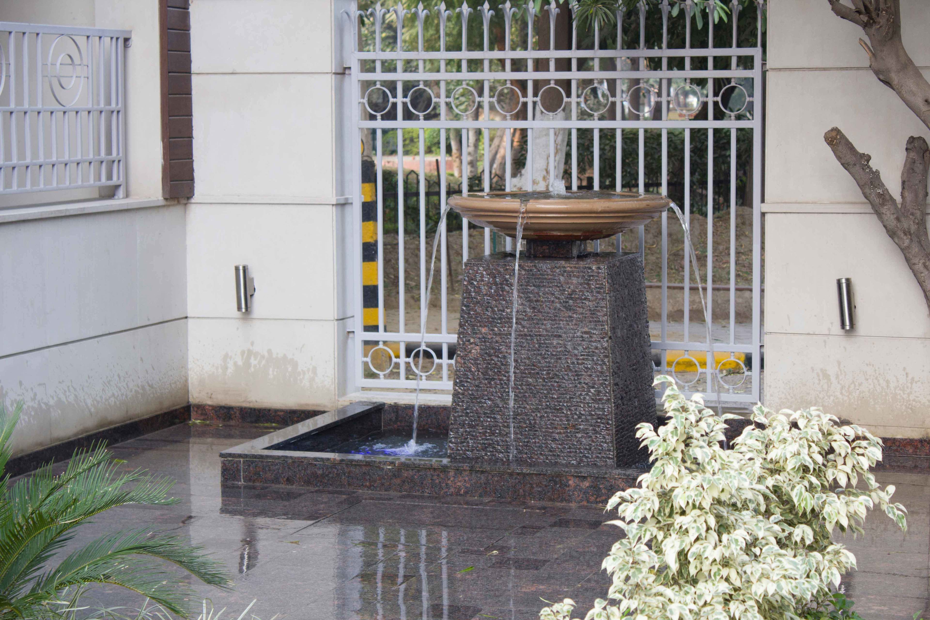 Une fontaine dont l’eau se déverse dans un bassin, le tout étant entouré d’une grille blanche et d’un arbre.