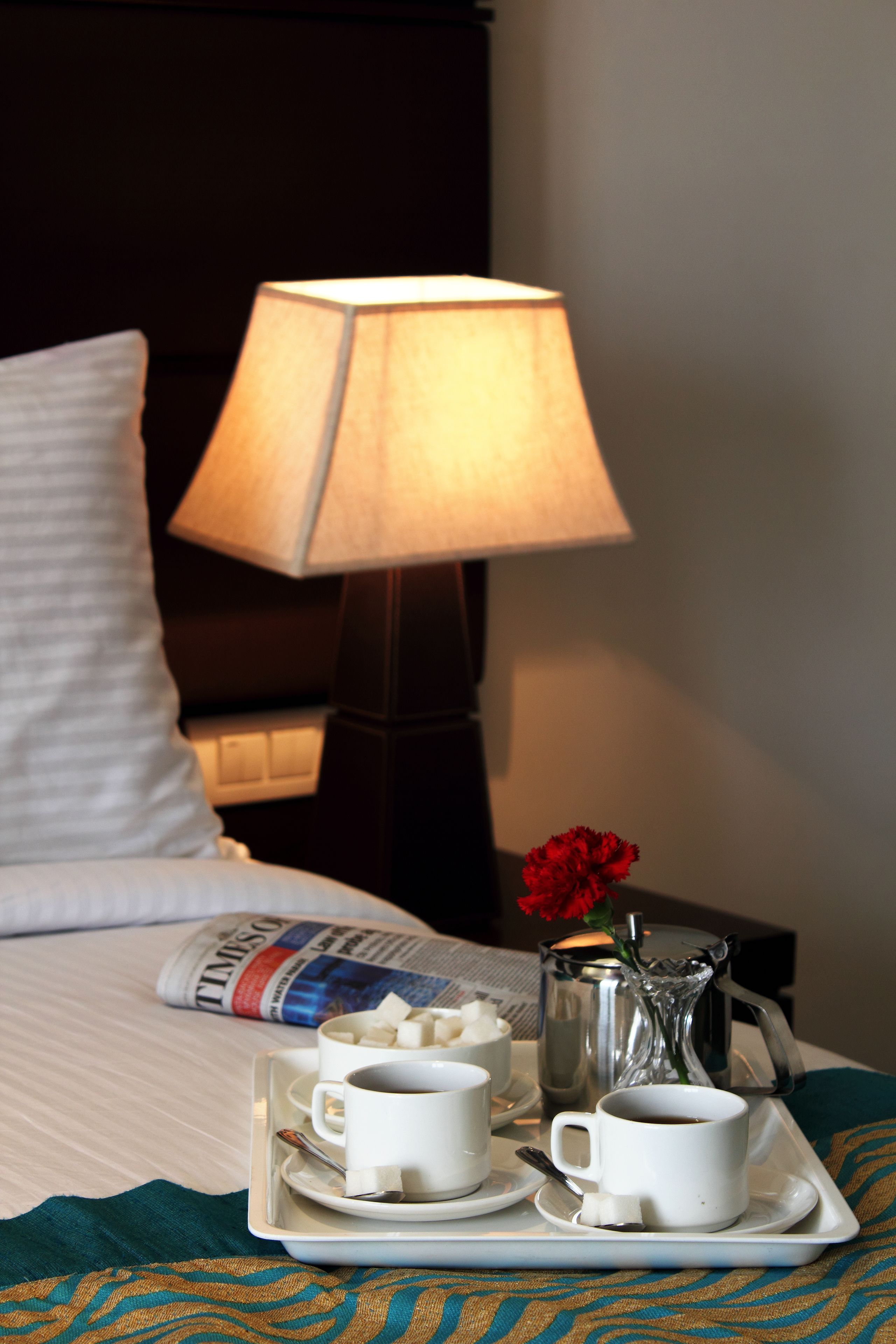 Ein ordentlich bezogenes Bett mit einem weißen Kopfkissen, ein Tablett mit zwei Tassen Kaffee und einem Teekessel, eine rote Blumenvase und eine Lampe auf einem Nachttisch.