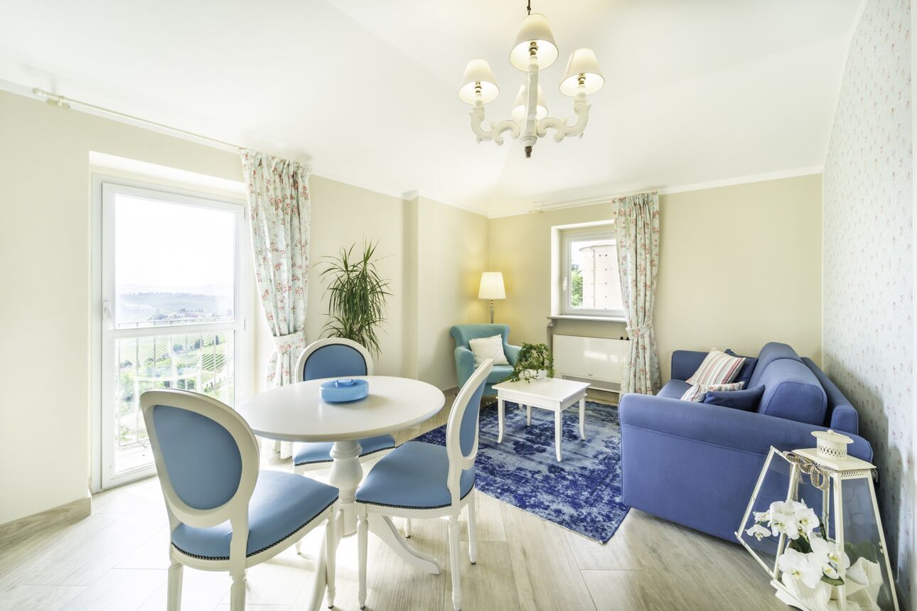 A living room with a blue sofa, a dining table with chairs, a chandelier, and a view through a large window.