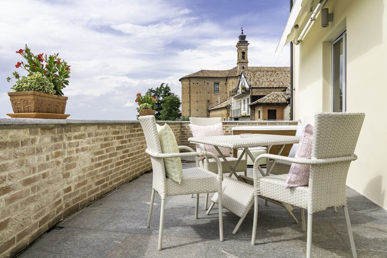 A balcony with wicker chairs and a table, a potted plant, and a building with a bell tower in the background.