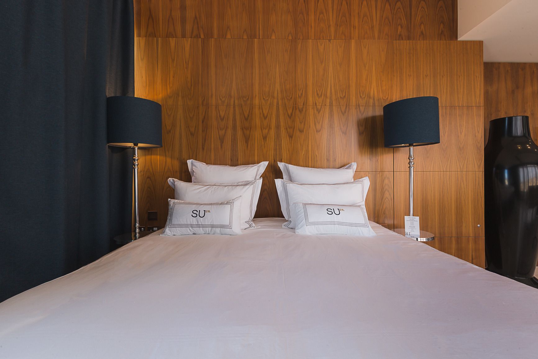 Ein Hotelzimmer mit einem hölzernen Kopfteil, zwei Nachttischlampen und einer großen Vase auf der rechten Seite.